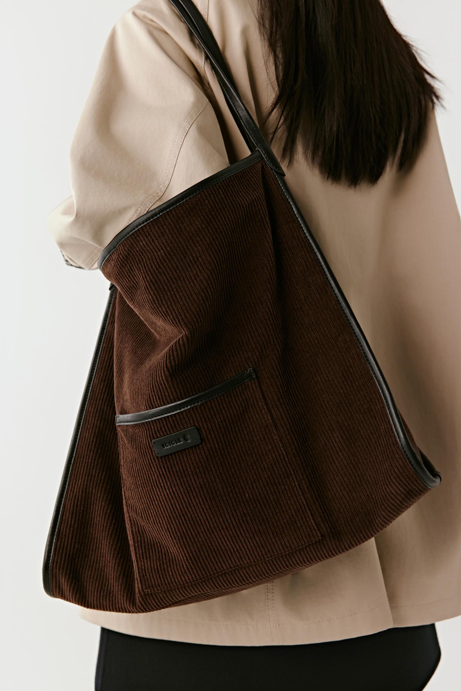 A dark brown corduroy tote bag with black trim and a shoulder strap, worn over the shoulder of a person wearing a beige blazer.