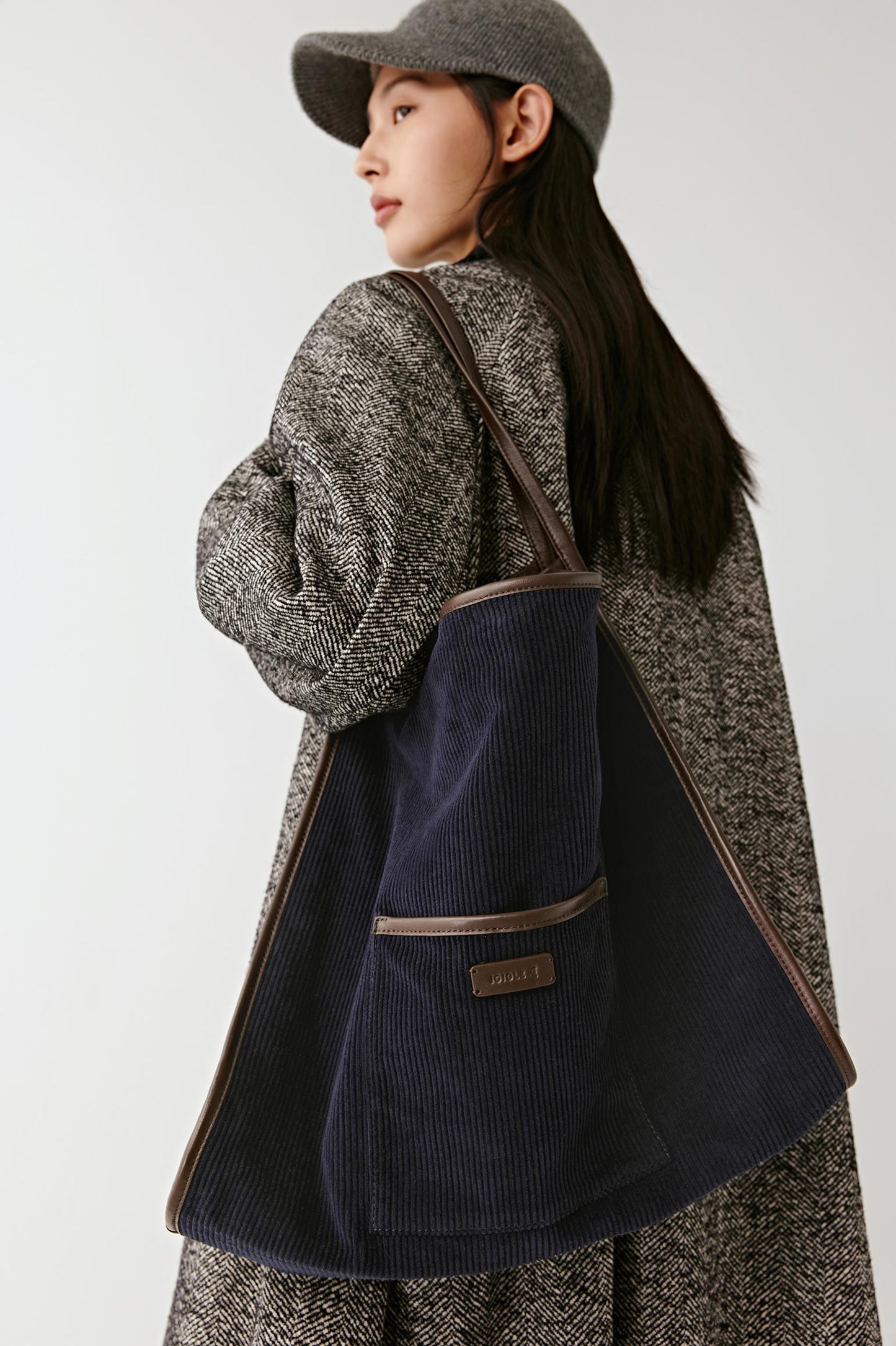 A woman wearing a blue textured coat and matching hat carries a large navy blue corduroy tote bag with brown trim and a small front patch logo, photographed against a plain light gray studio background.