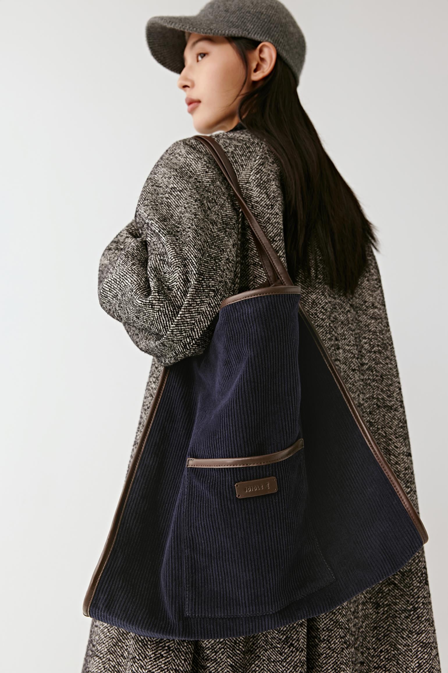 A woman wearing a blue textured coat and matching hat carries a large navy blue corduroy tote bag with brown trim and a small front patch logo, photographed against a plain light gray studio background.