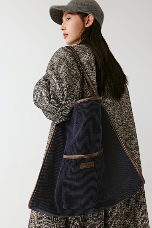 A woman wearing a blue textured coat and matching hat carries a large navy blue corduroy tote bag with brown trim and a small front patch logo, photographed against a plain light gray studio background.