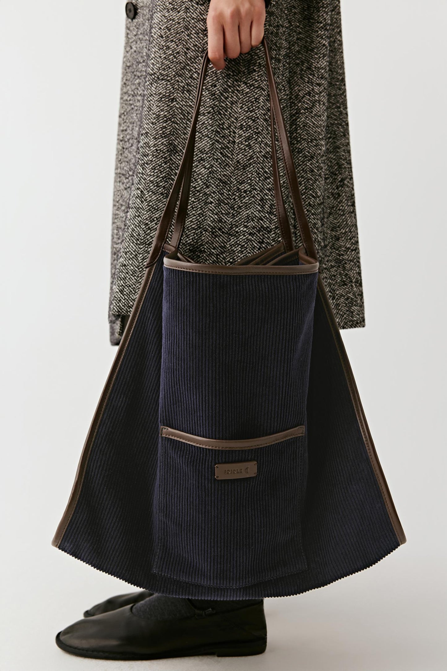 A person wearing a textured gray coat and black shoes holds a large navy corduroy tote bag with brown trim and shoulder straps against a light gray studio background.