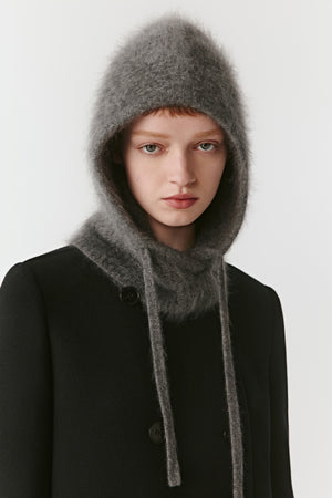 Woman wearing a grey knit hood with long drawstrings styled with a black coat on a light background
