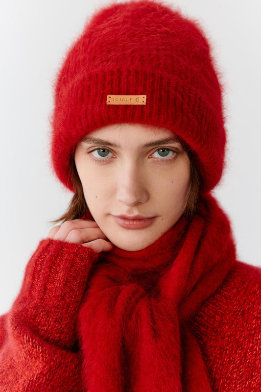 Portrait view of a woman wearing a deep red cashmere beanie styled with a matching scarf. The image highlights the plush texture, soft volume and fluffy surface of the cashmere knit, while the saturated red shade emphasizes the richness and depth of the material. A folded brim and contrasting front label complete the design.