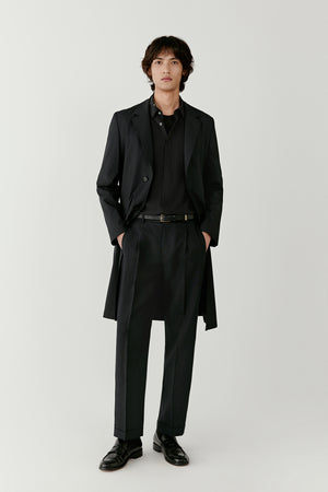 Front view of a male model wearing a straight-cut trench coat crafted from DEW wool and silk fabric in a dark grey melange shade. The coat features a clean silhouette with a tailored collar and minimal structure.