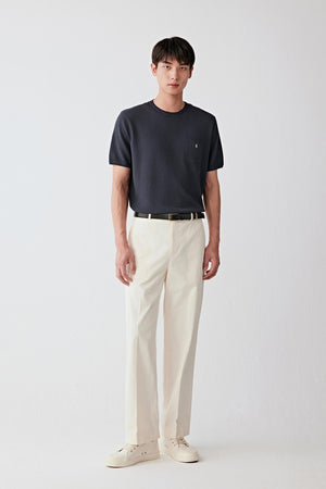 Full-length front view of a man standing facing forward wearing light beige cotton twill trousers paired with a dark T-shirt, highlighting the straight, relaxed silhouette, clean leg line and structured drape, with a smooth surface and compact weave characteristic of high-density cotton twill