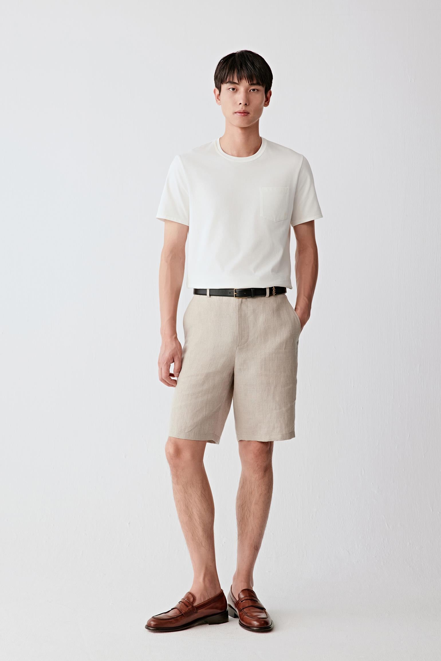 Front view of a man wearing beige tailored shorts in plain-woven linen, highlighting the straight silhouette, lightweight structure and naturally textured surface. The linen fabric reveals subtle irregularities in the weave, creating understated tonal variation across the garment.