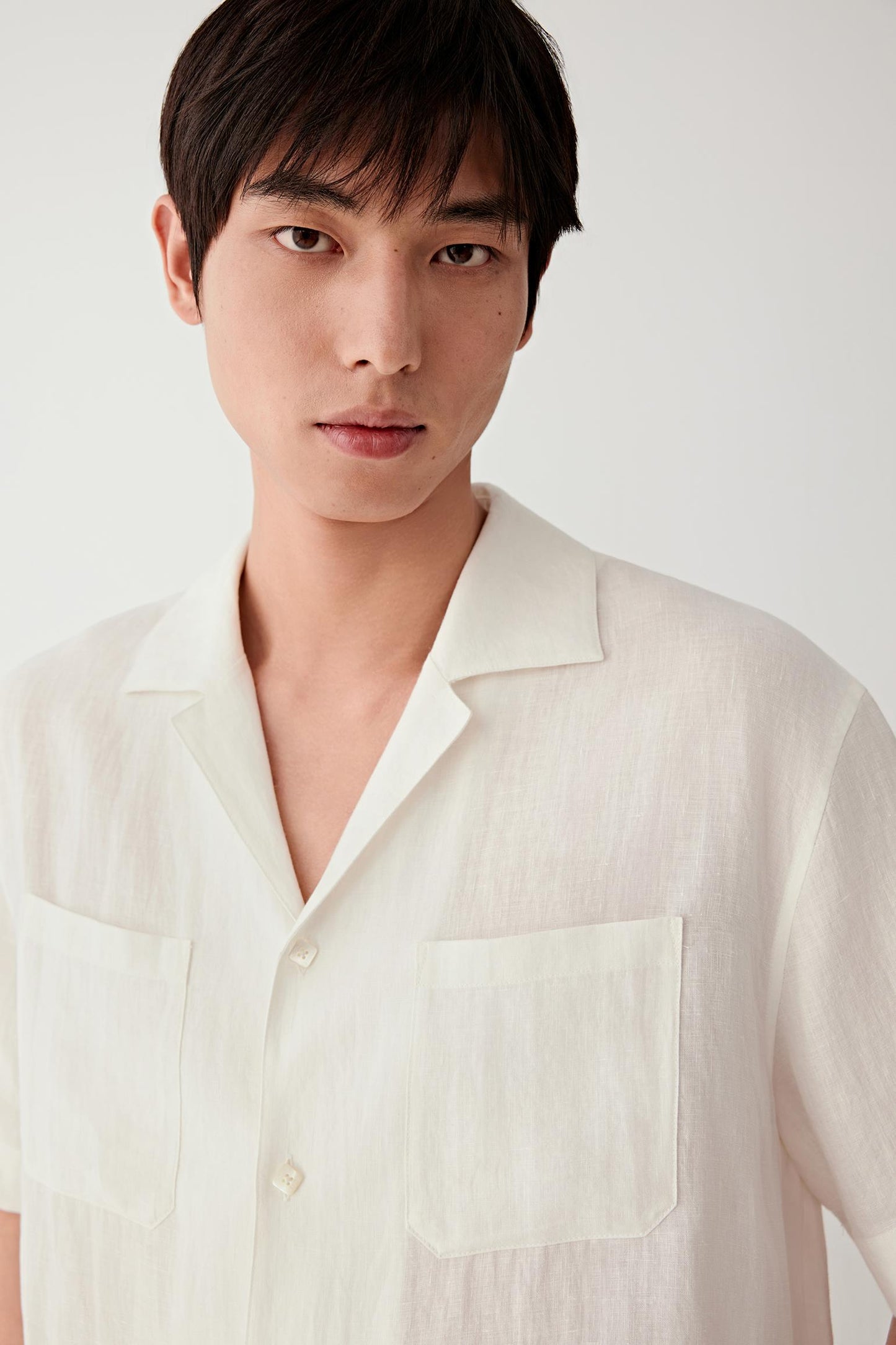Close-up portrait focusing on the open collar, front opening and dual chest pockets. The image reveals the dry hand, visible weave and understated texture characteristic of linen.