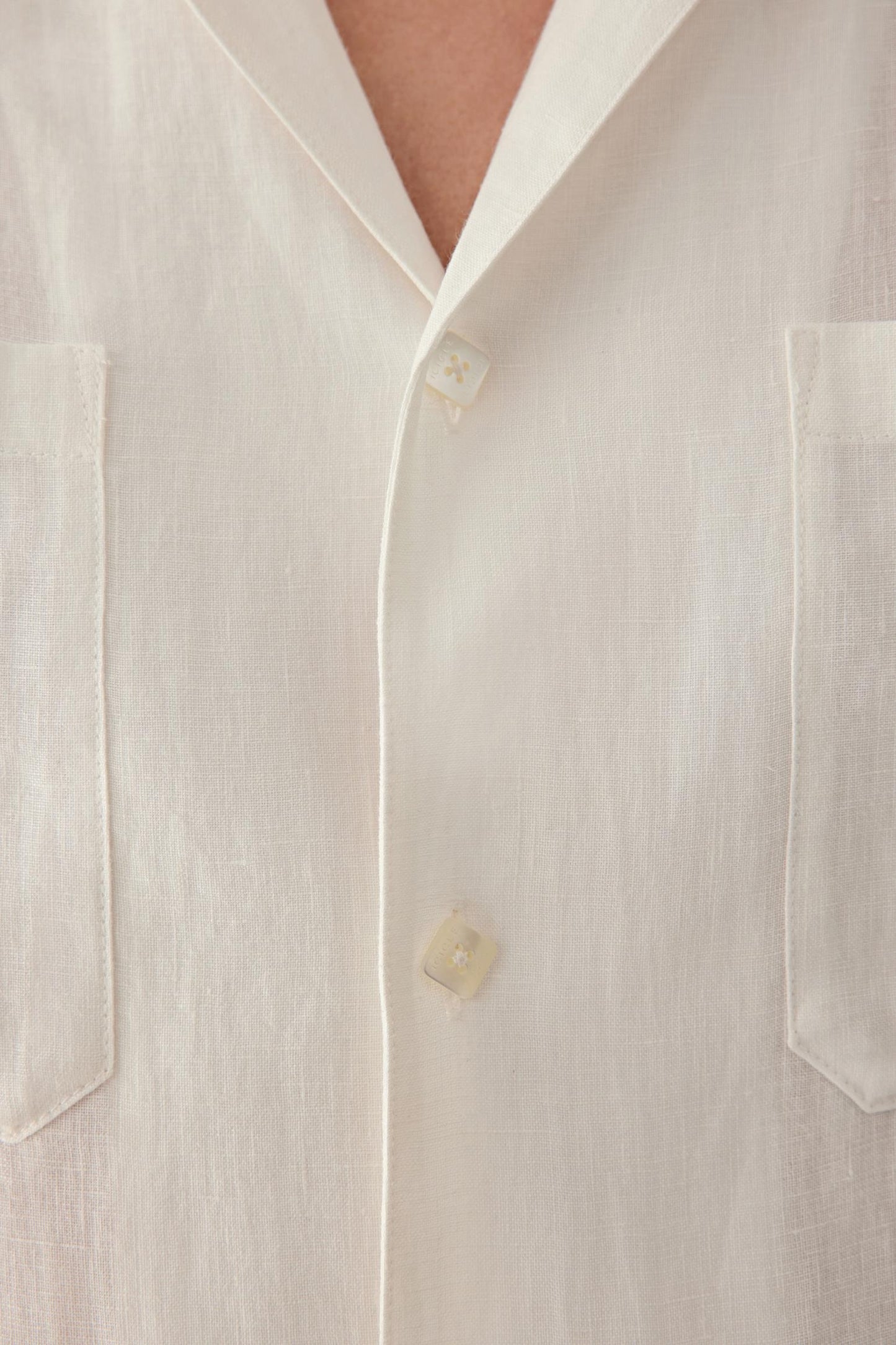 Close-up of the collar and front button fastening, showing the soft structure of the neckline, the natural texture of the linen surface and the subtle tonal depth created by the woven construction.