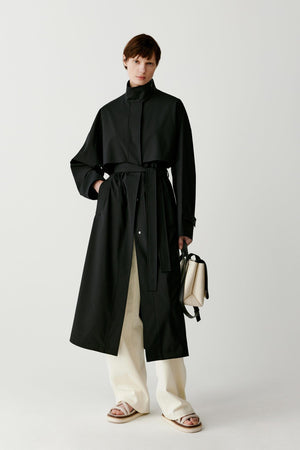 Full-length studio photograph of a woman standing against a light grey background wearing a long deep black single-breasted trench coat made from wool and silk. The coat has a straight elongated silhouette that falls below the calf, with a high structured collar, concealed front fastening and long sleeves. Her left hand rests in the coat pocket while she holds a light-coloured handbag in the other hand. The coat is worn over light trousers and beige shoes.