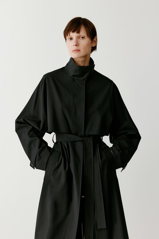 Front view of the long black trench coat showing the straight silhouette, long sleeves and waist belt tied at the front, defining the coat’s shape while keeping a fluid drape.