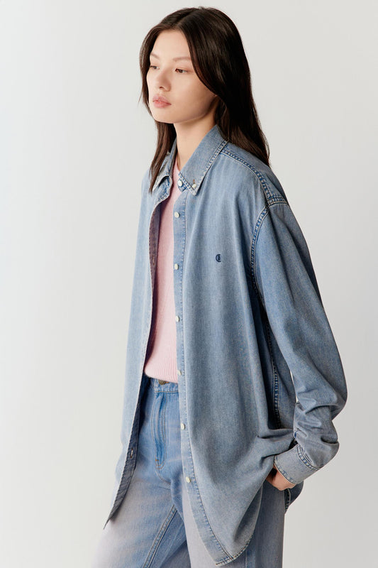 Three-quarter front view of a woman wearing a light blue cotton denim shirt with a soft washed finish and subtle pinkish undertones, featuring a relaxed straight cut, classic collar, long sleeves and fluid drape, styled with matching denim trousers, highlighting the lightweight structure and natural fall of the fabric