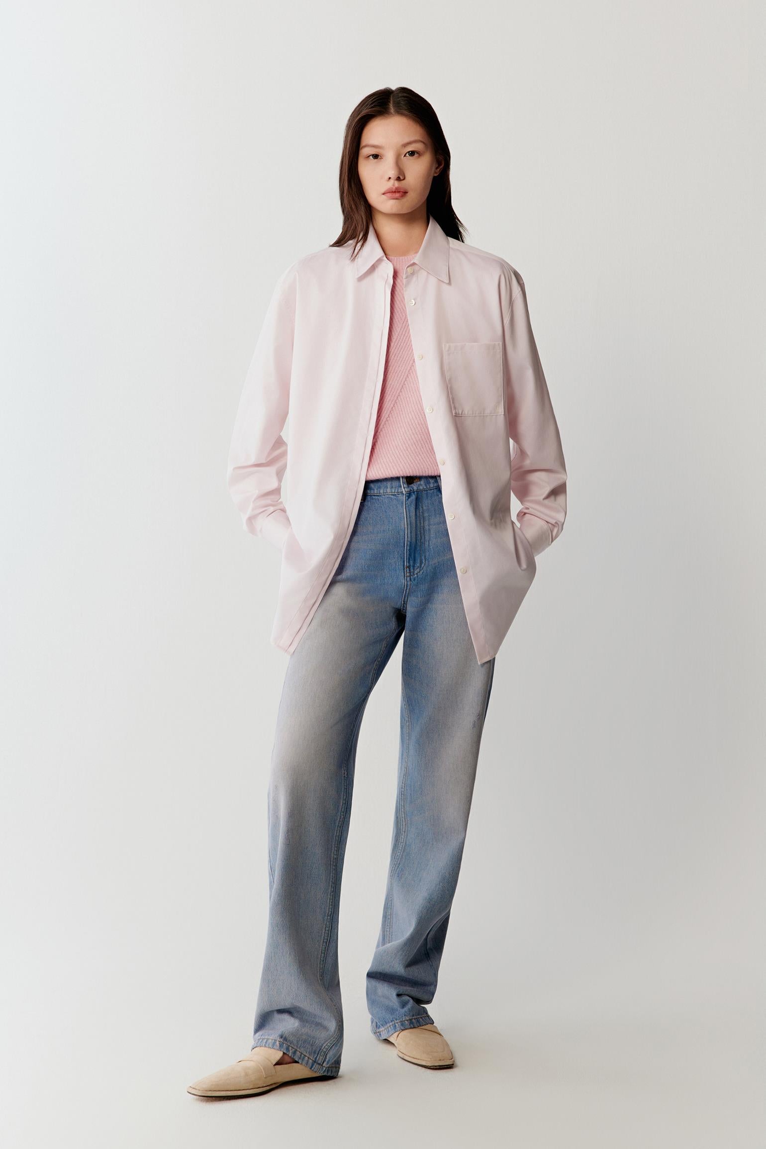 Full-length studio photograph of a woman standing against a light grey background wearing a light pink striped shirt made from yarn-dyed cotton twill. The shirt has a relaxed straight silhouette with a pointed collar, long sleeves and a front button placket. It is worn with blue denim jeans.