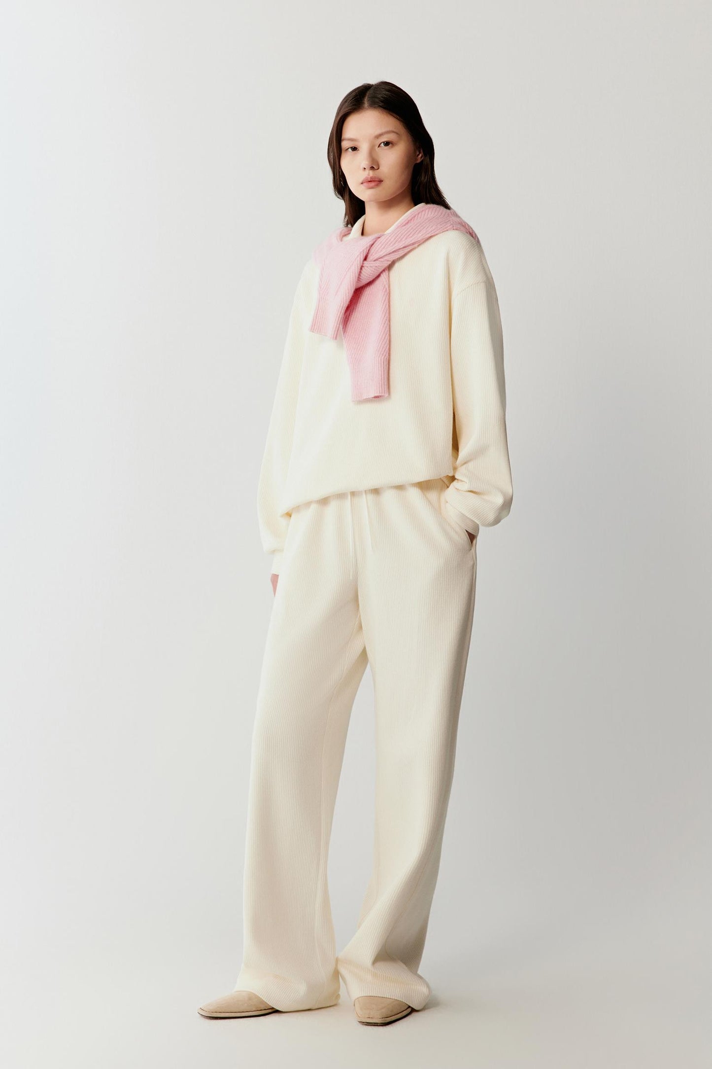 Full-length front view of a model wearing a cream sweater crafted from stretch rib jersey. The sweater features a relaxed silhouette with a soft open collar and long sleeves. The lightweight ribbed structure creates vertical texture while maintaining fluid movement.