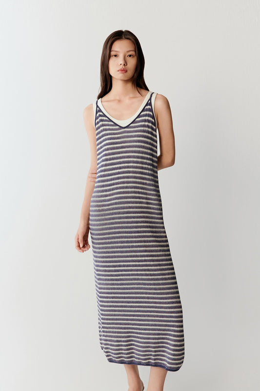 Full-length front view of a woman wearing a knitted linen and silk striped dress with thin navy, off-white and subtle red stripes, highlighting the fluid straight silhouette and lightweight knit texture