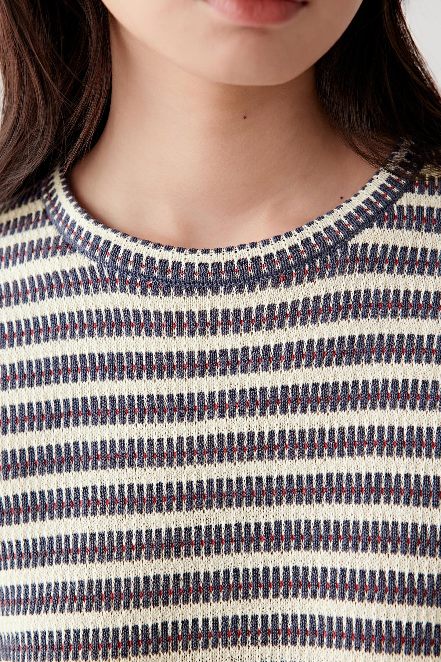 Close-up portrait focusing on the round neckline and striped knit surface, detailing the alternating navy, off-white and muted red yarns with a lightly textured appearance