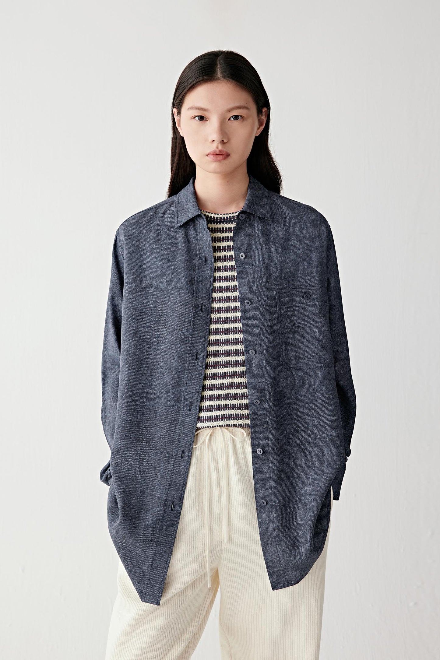 Mid-length front view of a woman wearing an indigo blue silk twill shirt layered over a striped knit top and paired with ivory trousers, highlighting the fluid straight silhouette, lightweight texture and softly structured drape of the silk twill fabric