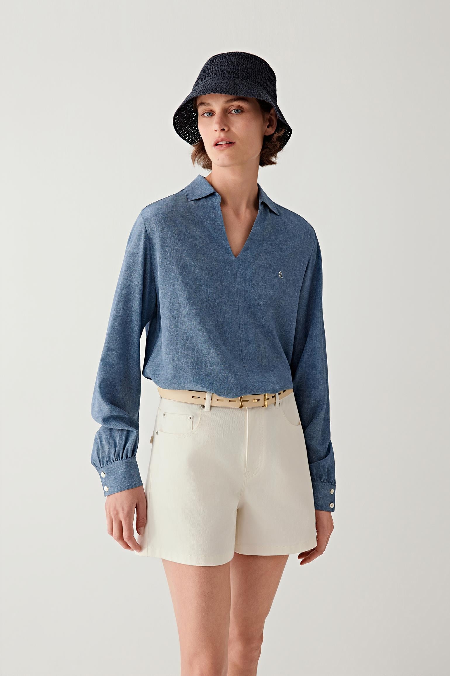 Full-length front view of a woman standing facing forward wearing a light blue shirt and off-white shorts, styled with a slim light beige calf leather belt at the waist, showing the narrow width, smooth leather surface and how the belt subtly structures the silhouette with a clean, minimal line