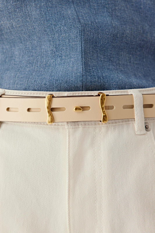 Close-up view of the waist focusing on the belt worn over light-coloured trousers, highlighting the elongated perforations, the precise alignment of the holes, the smooth calf leather finish and the antique gold-tone brass buckle positioned at the front