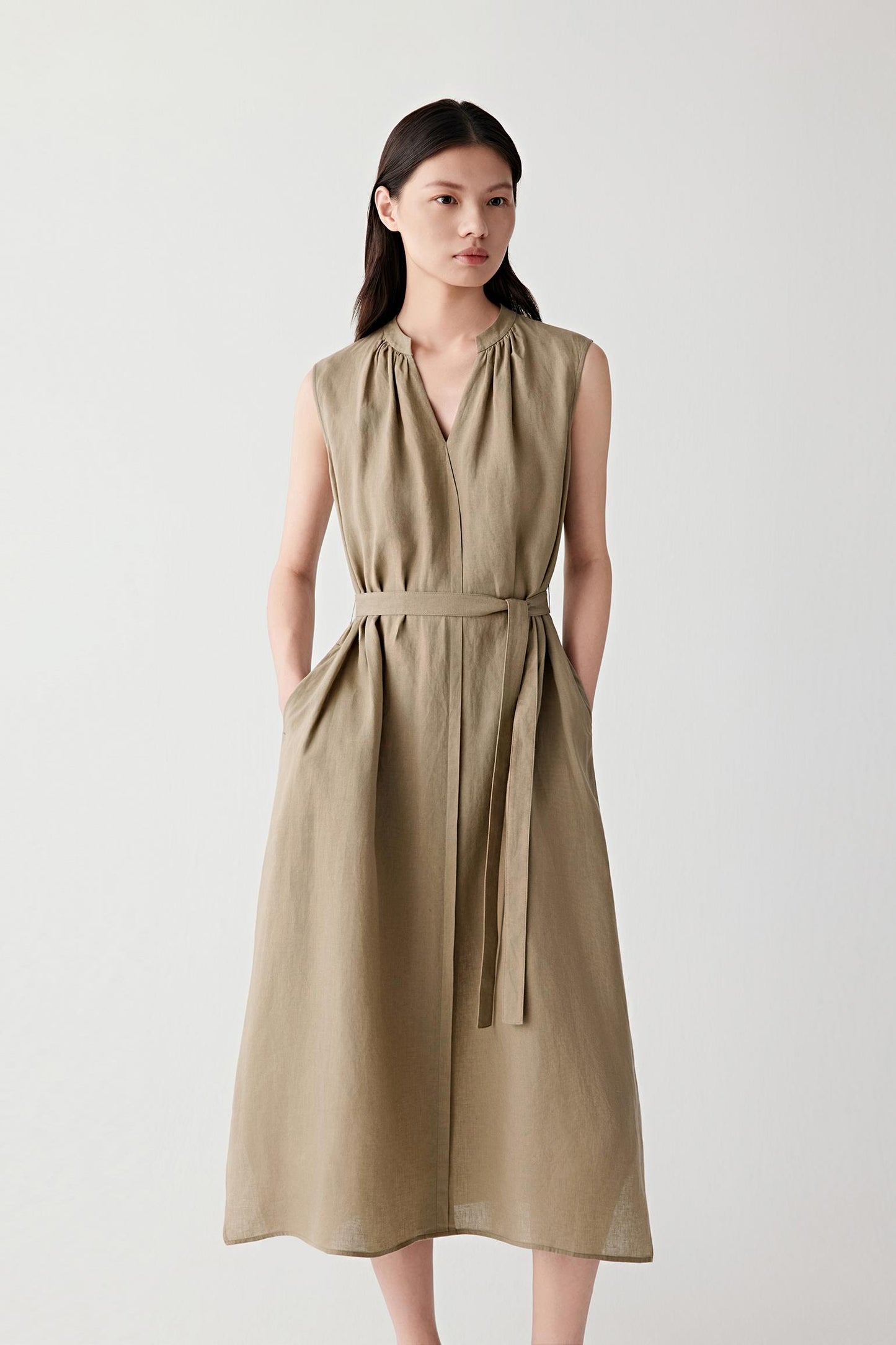 Front view of a woman wearing a brown linen dress, highlighting the fluid silhouette, visible grain and naturally textured surface of the fabric. The sleeveless construction, gathered V-shaped neckline and self-tie waist belt create a balanced shape with soft structure and movement.