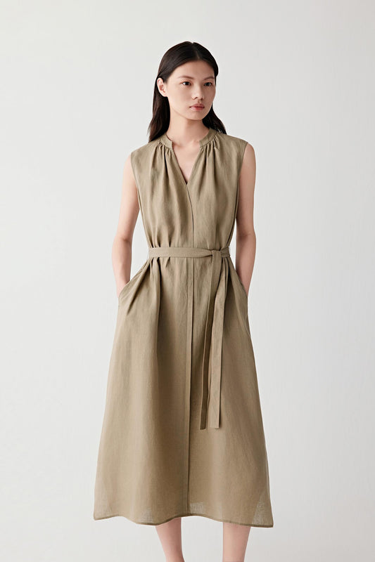 Front view of a woman wearing a brown linen dress, highlighting the fluid silhouette, visible grain and naturally textured surface of the fabric. The sleeveless construction, gathered V-shaped neckline and self-tie waist belt create a balanced shape with soft structure and movement.