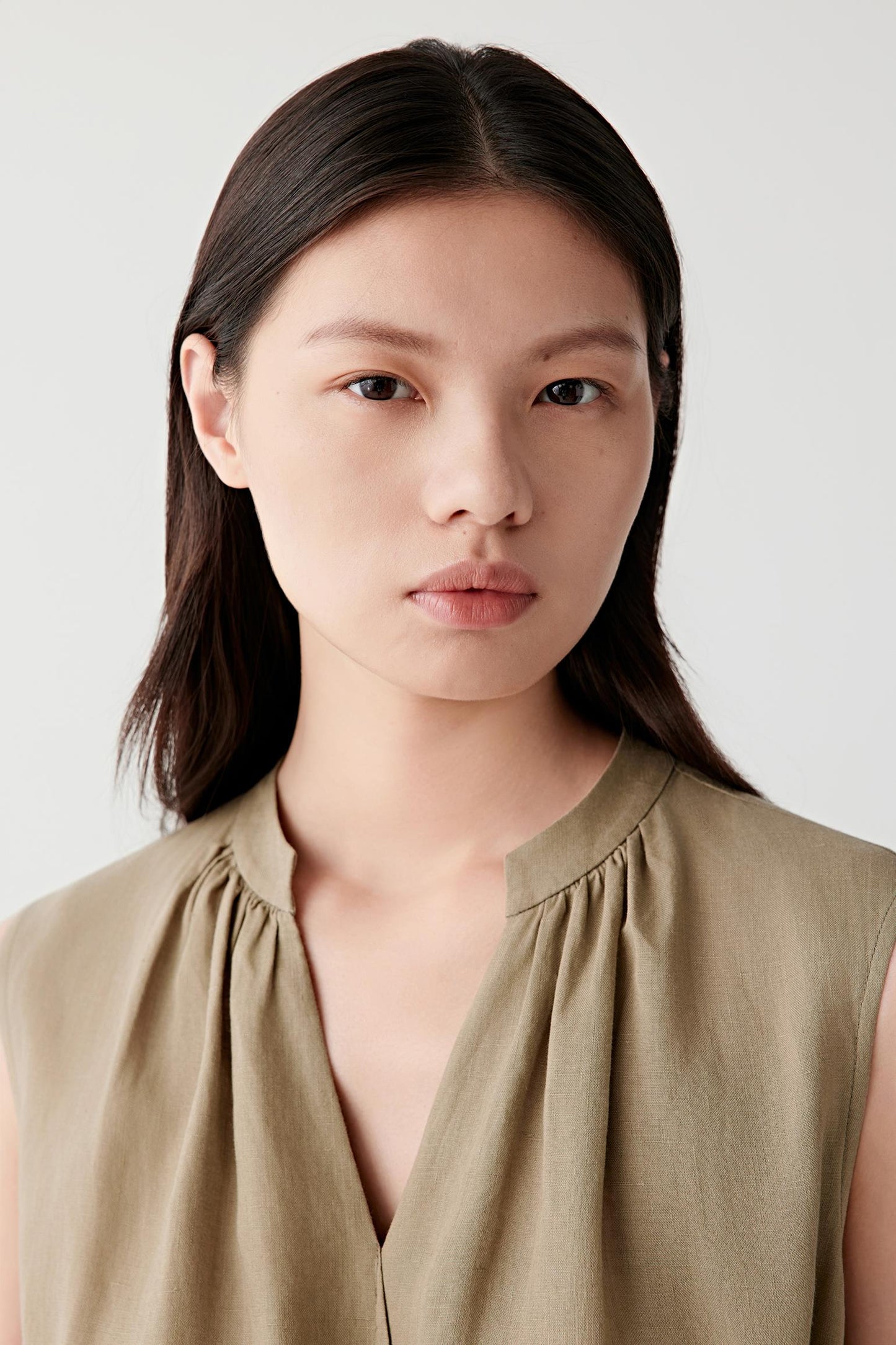 Close-up portrait focusing on the gathered V-shaped neckline and upper body construction. The image reveals the dry hand, visible weave and natural tonal variation of the linen fabric while highlighting the refined detailing around the collar area.