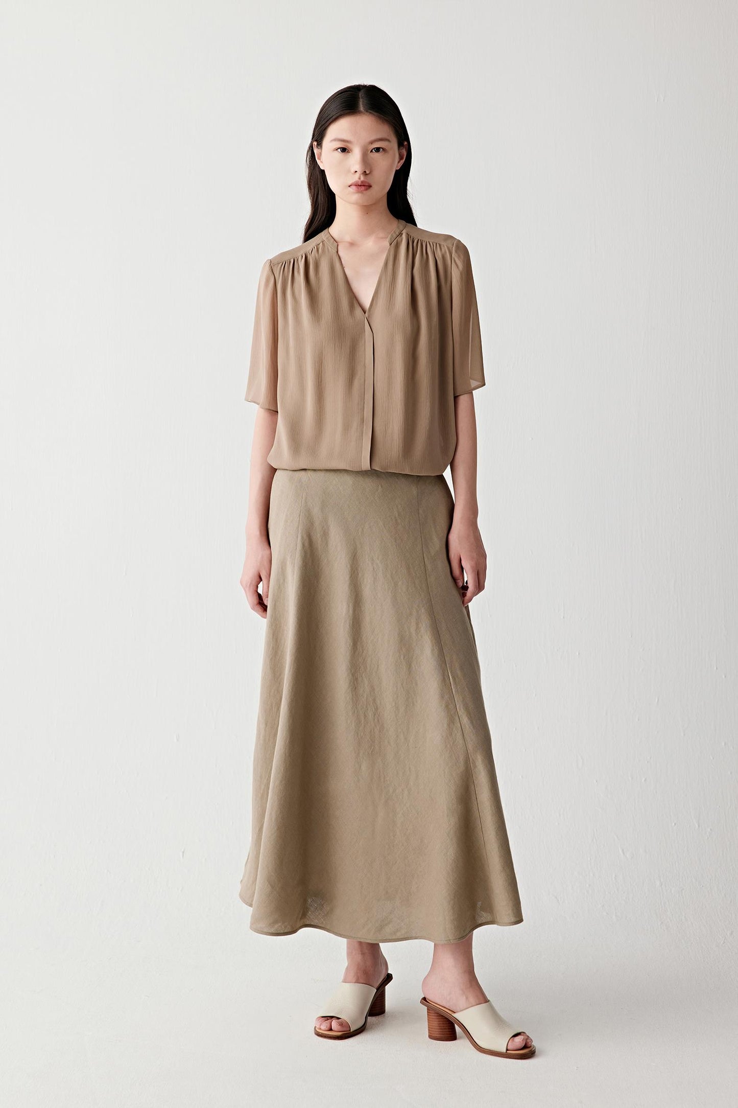 Front view of a woman wearing a taupe linen skirt, highlighting the fluid silhouette, lightweight structure and naturally textured surface. The visible grain of the linen fabric creates subtle depth and tonal variation across the garment.