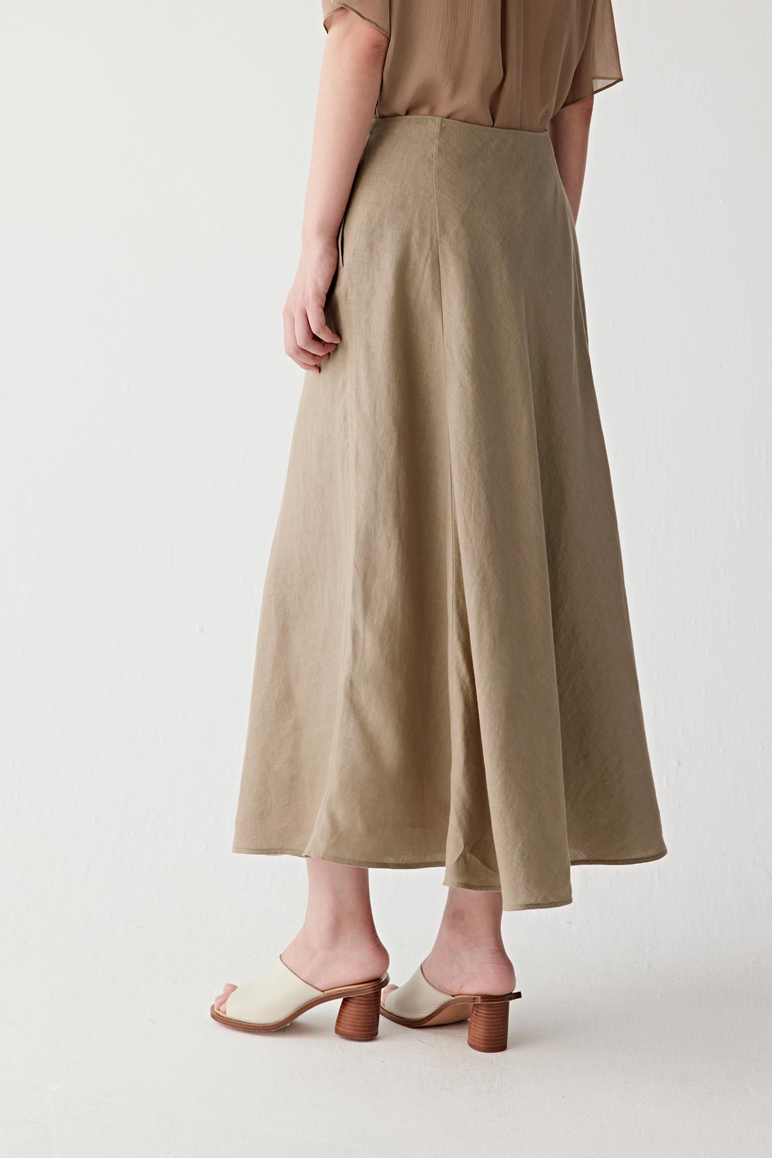 Angled rear view revealing the volume and shape created by the panelled construction. The image emphasizes the continuous drape of the linen fabric and the elegant flow of the flared hem.