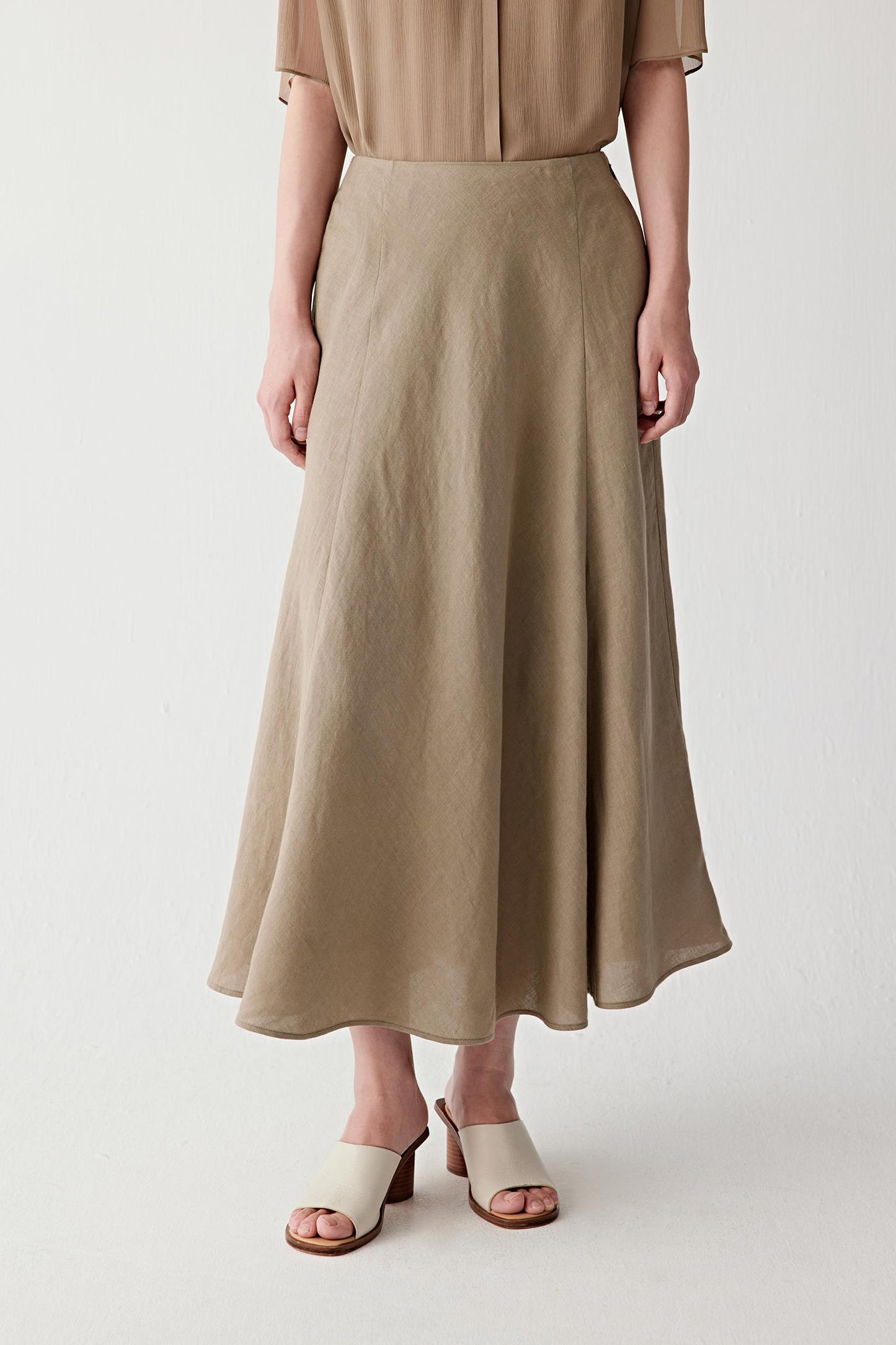 Three-quarter view showing the long flared silhouette and the soft movement of the linen fabric. The image emphasizes the balanced proportions, fluid drape and understated character of the natural fibre.