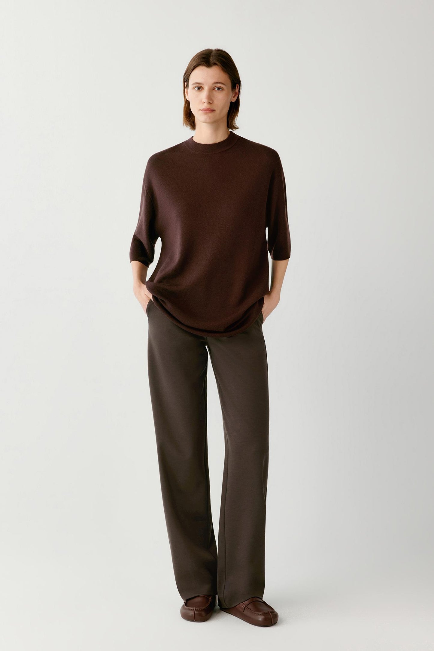 A woman stands against a light gray studio background wearing a dark brown long-sleeve knit top and matching dark brown wide-leg trousers, with brown shoes.