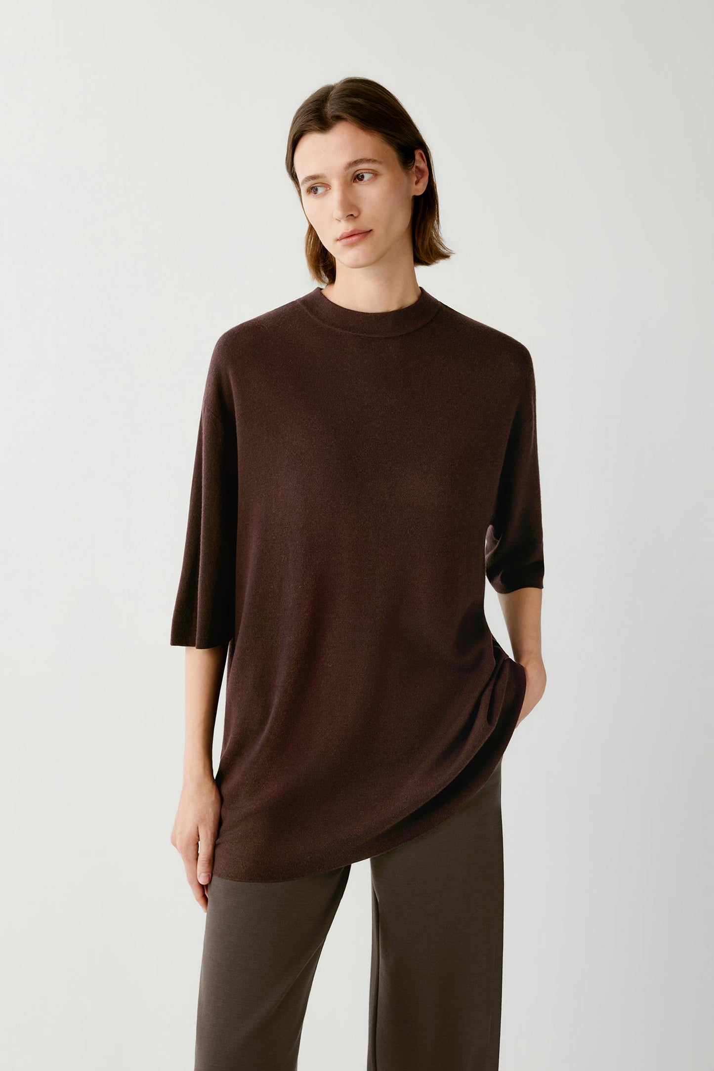 A woman modeling a dark brown knit top with three-quarter sleeves and a relaxed fit, paired with dark brown trousers, against a light gray studio background.