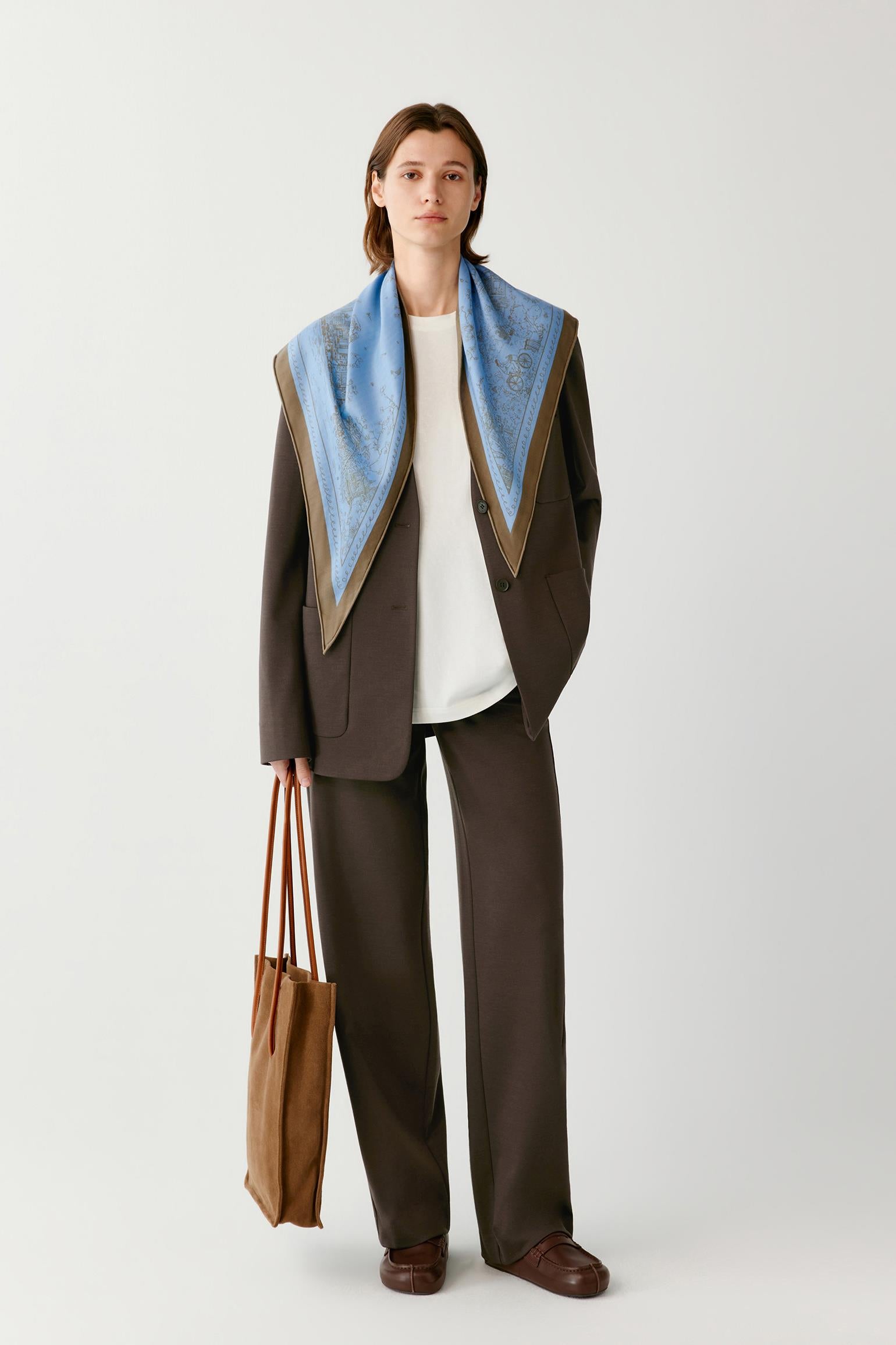Woman wearing a brown suit with a blue patterned scarf, carrying a brown tote bag.