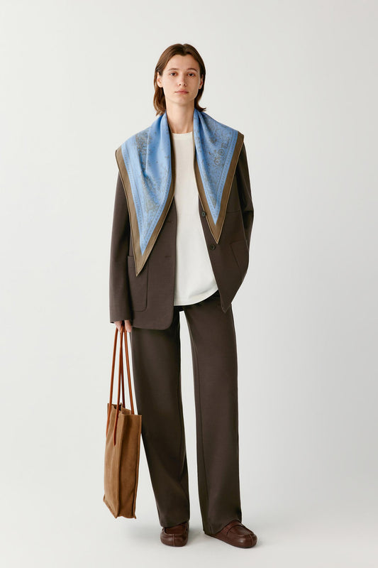 Woman wearing a brown suit with a blue patterned scarf, carrying a brown tote bag.
