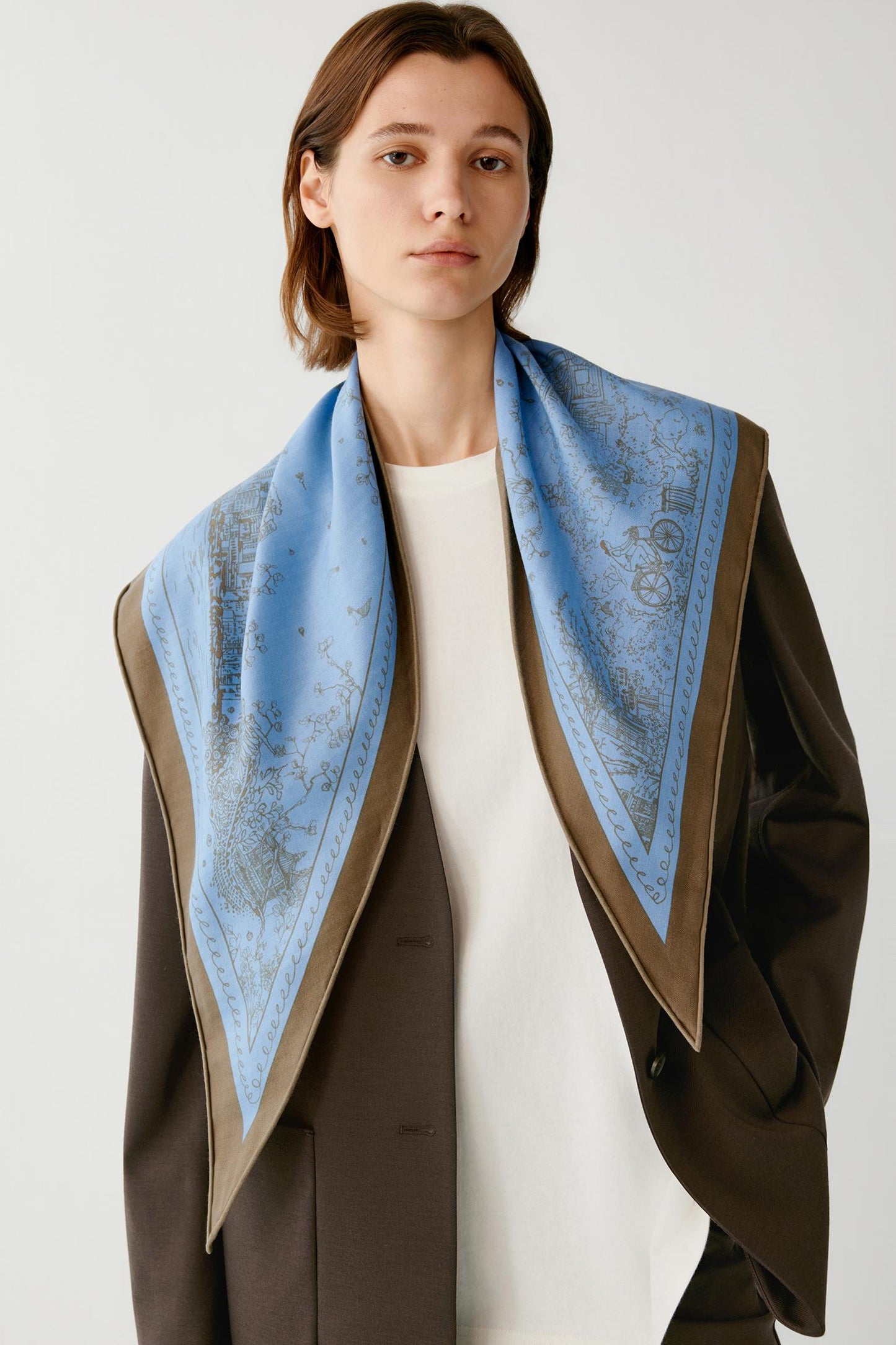 Woman wearing a blue patterned scarf over a brown blazer and white top.