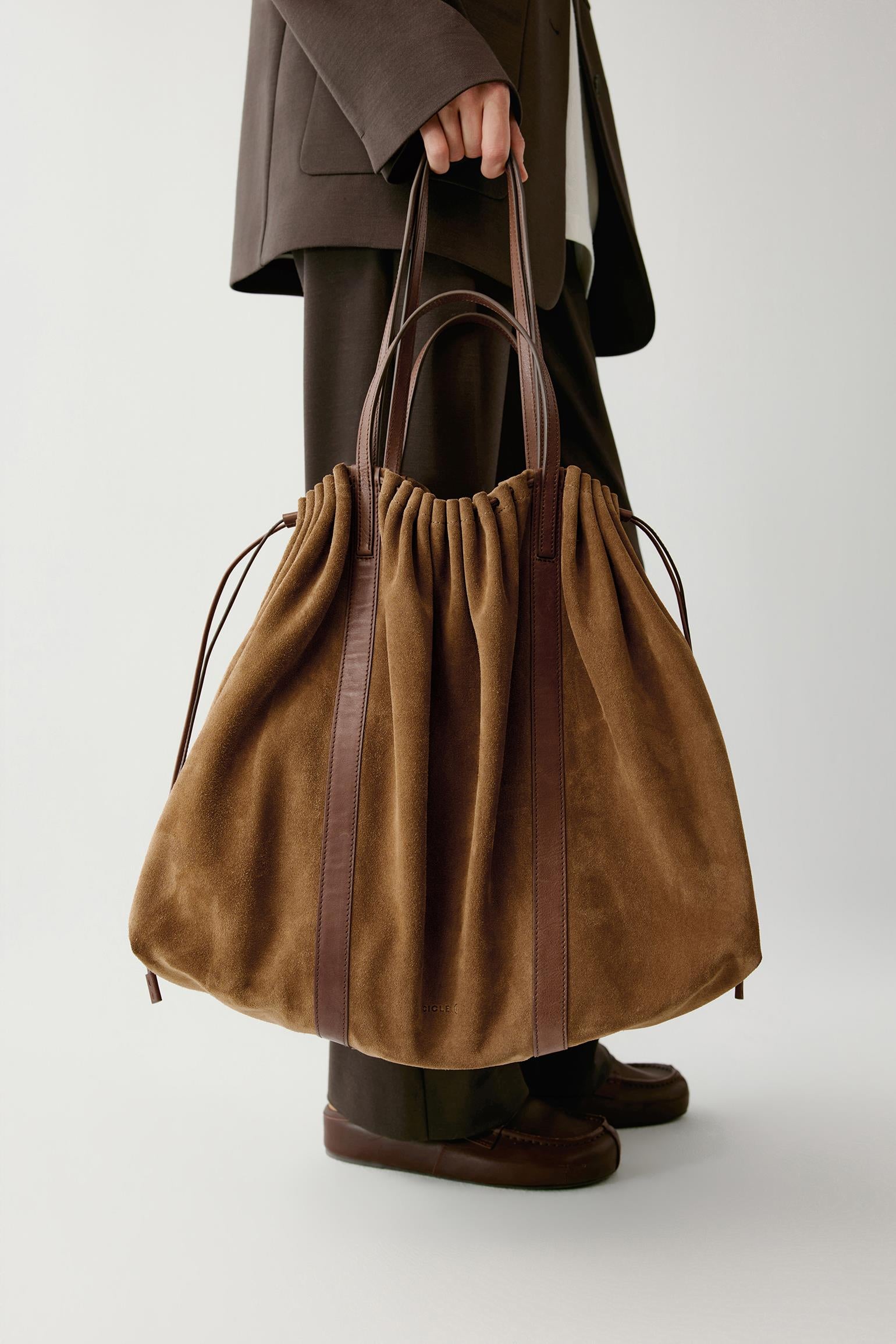 A person holds a large brown suede drawstring bag with dark brown leather handles and vertical trim against a light gray studio background.