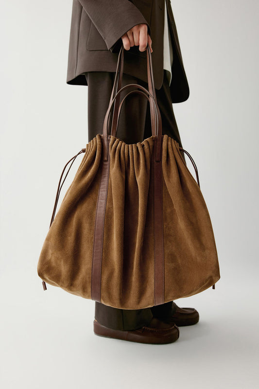 A person holds a large brown suede drawstring bag with dark brown leather handles and vertical trim against a light gray studio background.