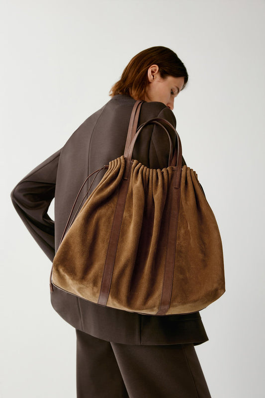 A model wearing a dark brown blazer and matching trousers carries a large brown suede drawstring tote bag over their shoulder, photographed against a neutral light gray studio background.