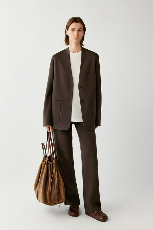 Woman wearing a brown collarless suit over a white top, carrying a large brown bag.