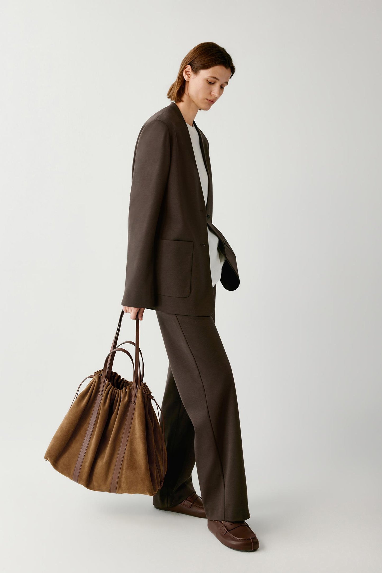 A model in a dark brown tailored suit holding a large brown leather tote bag, photographed against a clean light gray studio background.