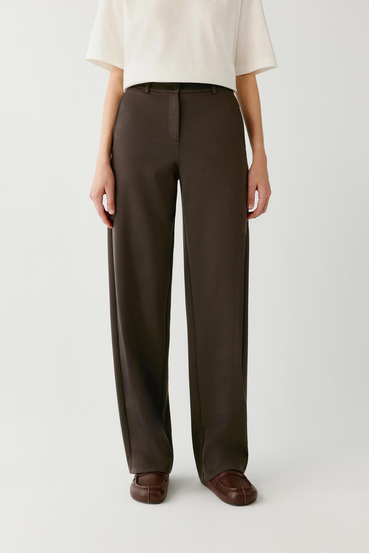 Woman wearing brown straight-leg tailored trousers with a white top and brown loafers.