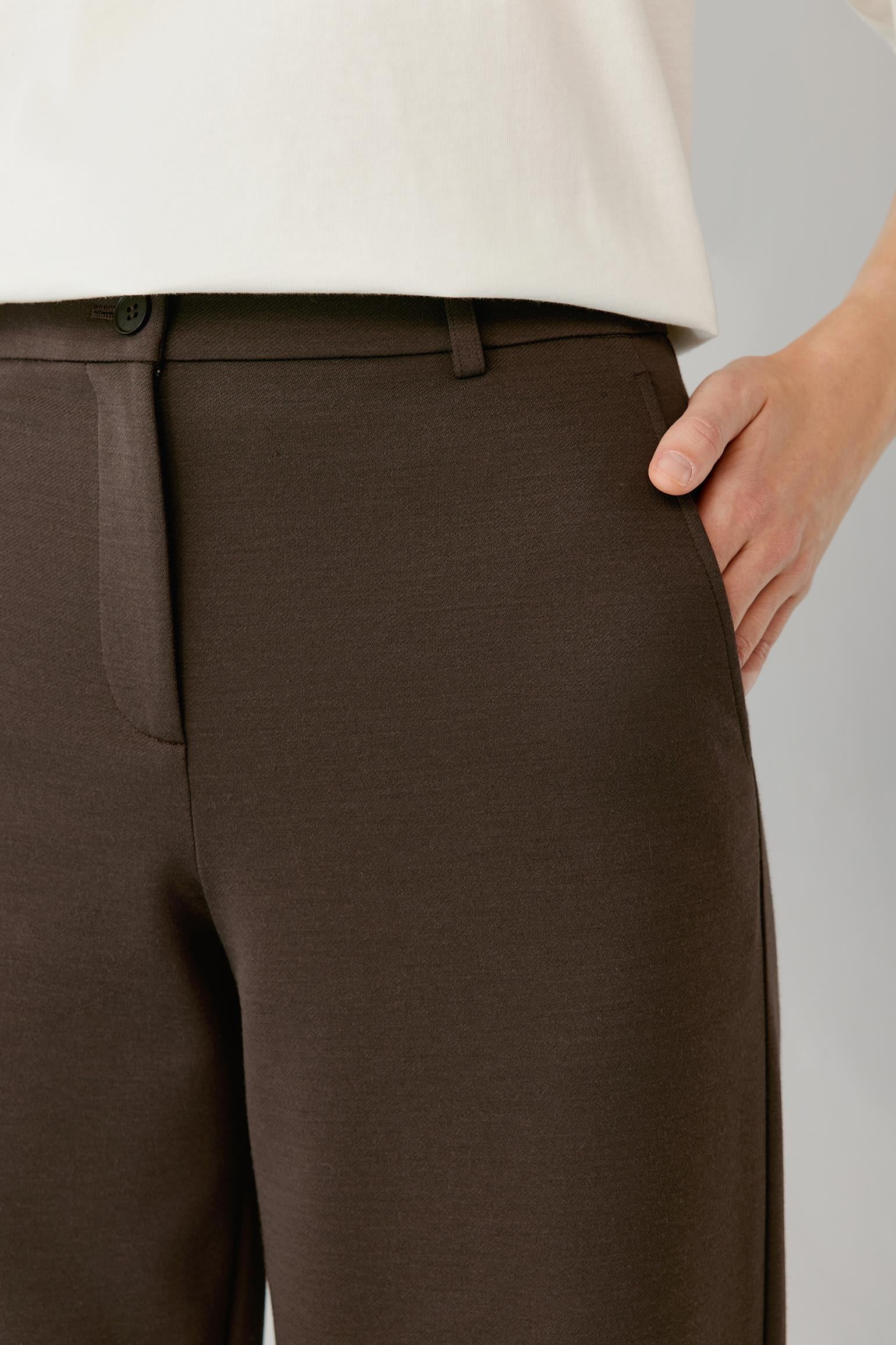 Close-up of the waistband, front closure and side pocket of brown tailored trousers.