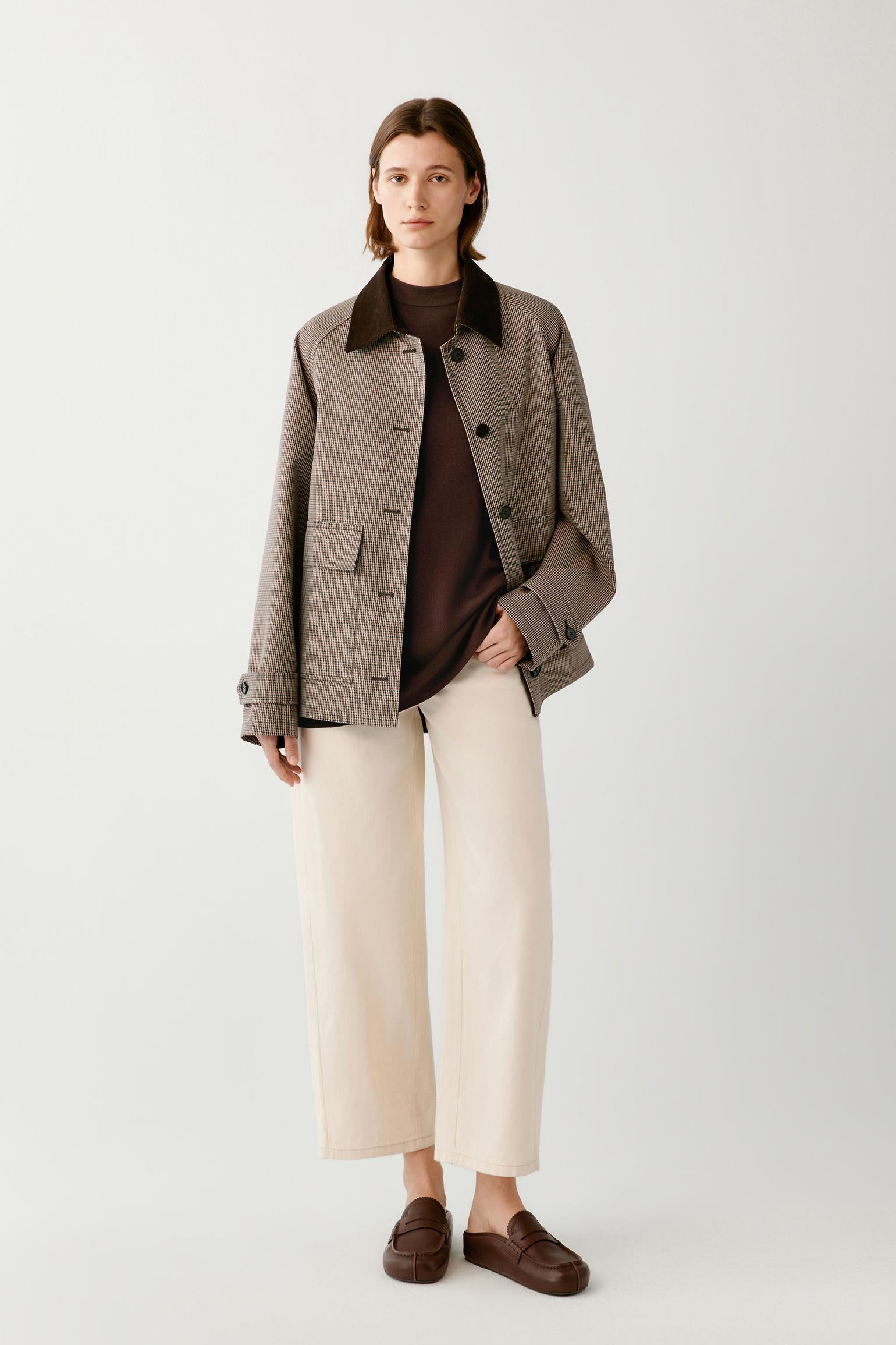 A model stands against a light gray studio background wearing a taupe-gray textured jacket with a dark brown corduroy collar, a dark brown turtleneck, cream wide-leg cropped trousers, and dark brown loafers.