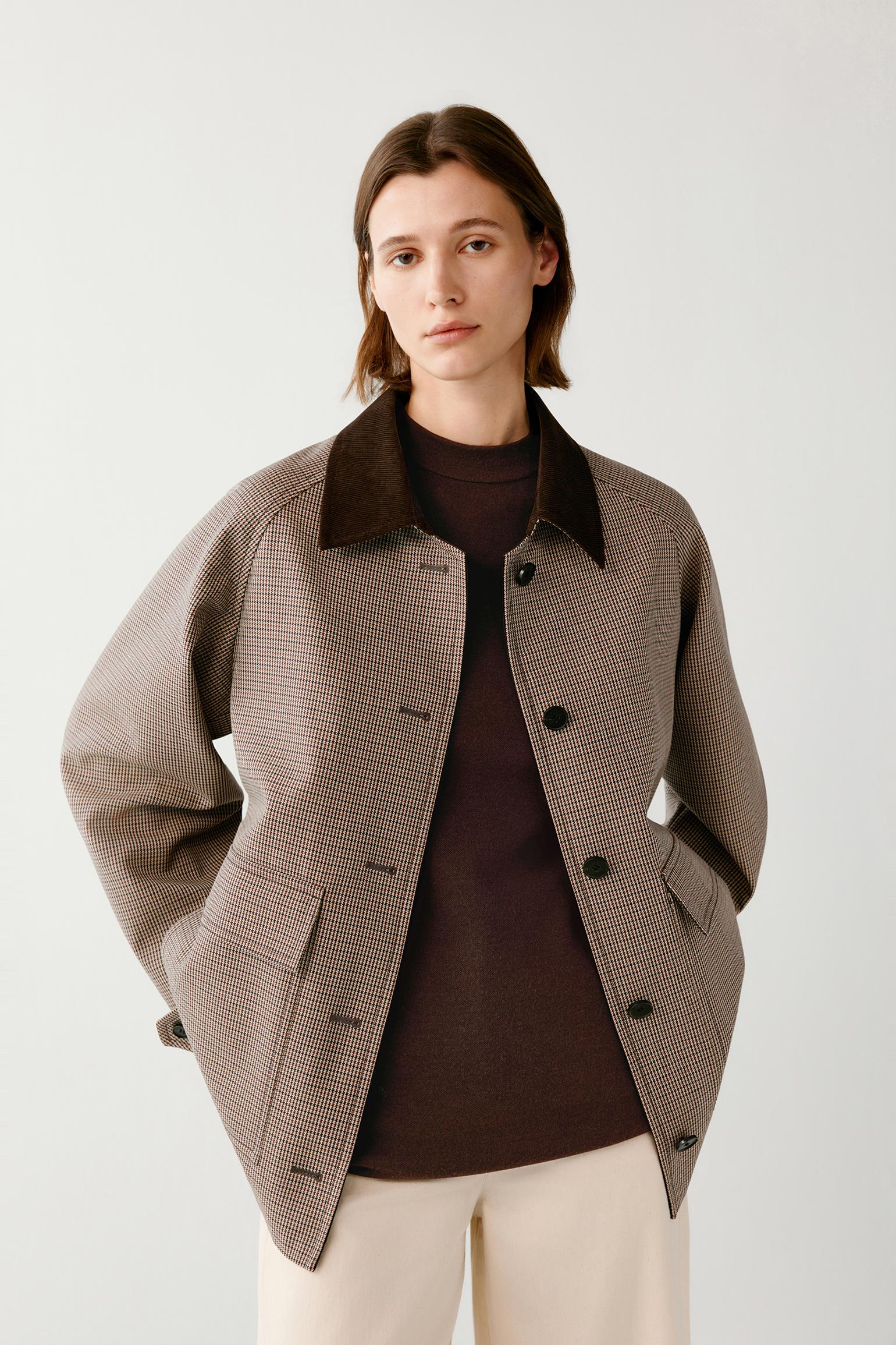 A woman modeling a brown and beige checkered jacket with a dark brown corduroy collar, worn over a dark brown top and cream trousers, against a light gray studio background.