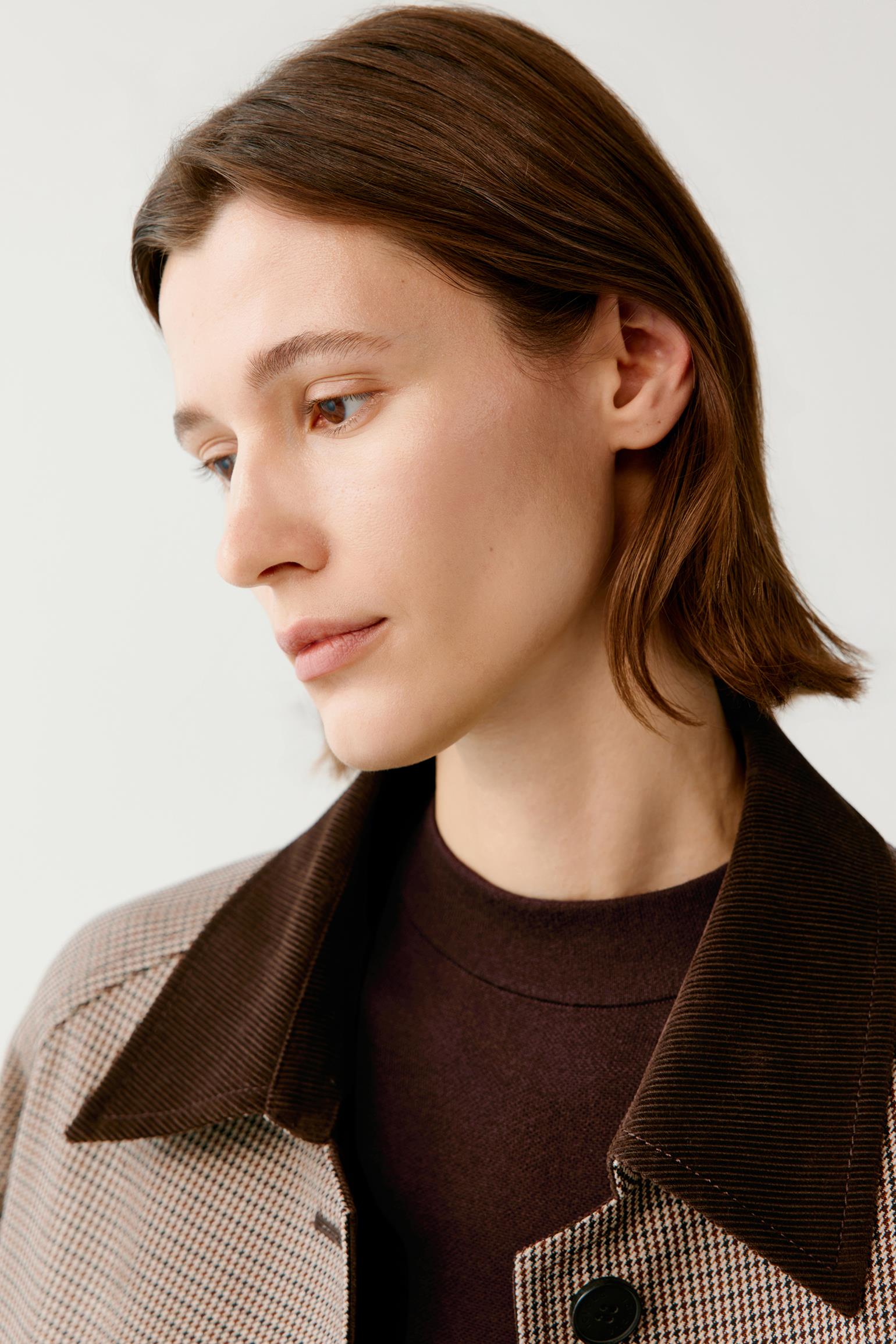 A close-up of a woman wearing a brown corduroy collar jacket over a dark brown sweater, set against a plain light gray background.