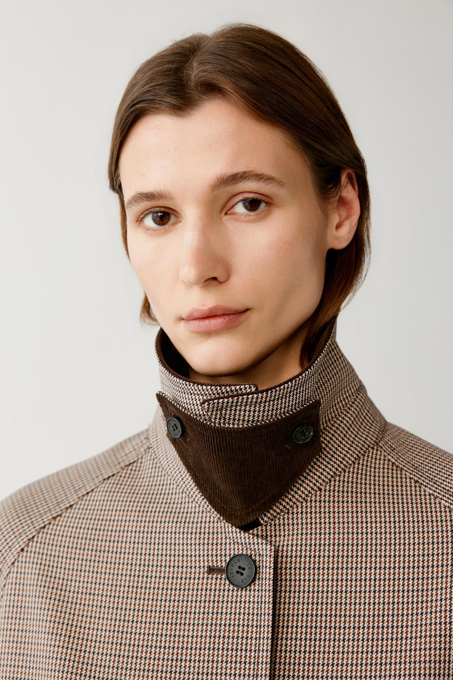A close-up portrait of a model wearing a brown and beige houndstooth patterned jacket with a dark brown corduroy high collar.