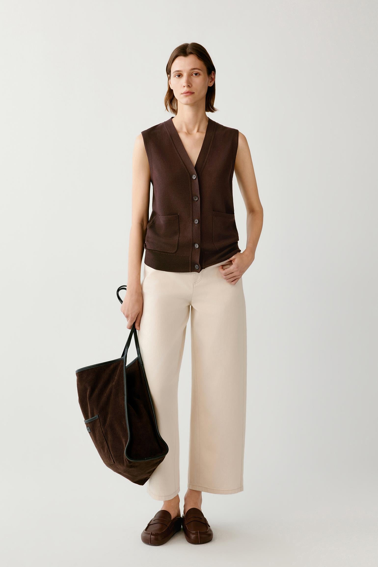 A model stands against a light gray studio background wearing a dark brown sleeveless button-front vest, cream wide-leg cropped pants, and brown slip-on shoes, while holding a large dark brown tote bag.