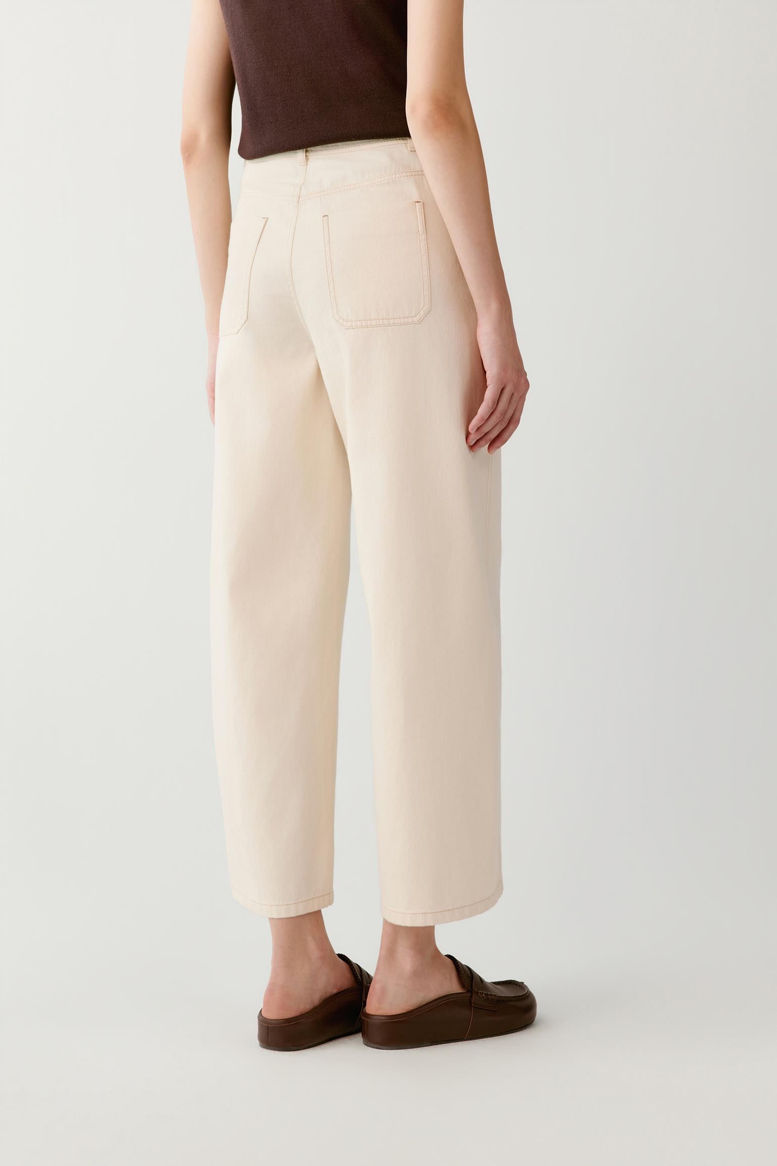 A person shown from the back wearing cream-colored cropped wide-leg trousers, a dark brown sleeveless top, and dark brown slip-on shoes against a plain light gray studio background.