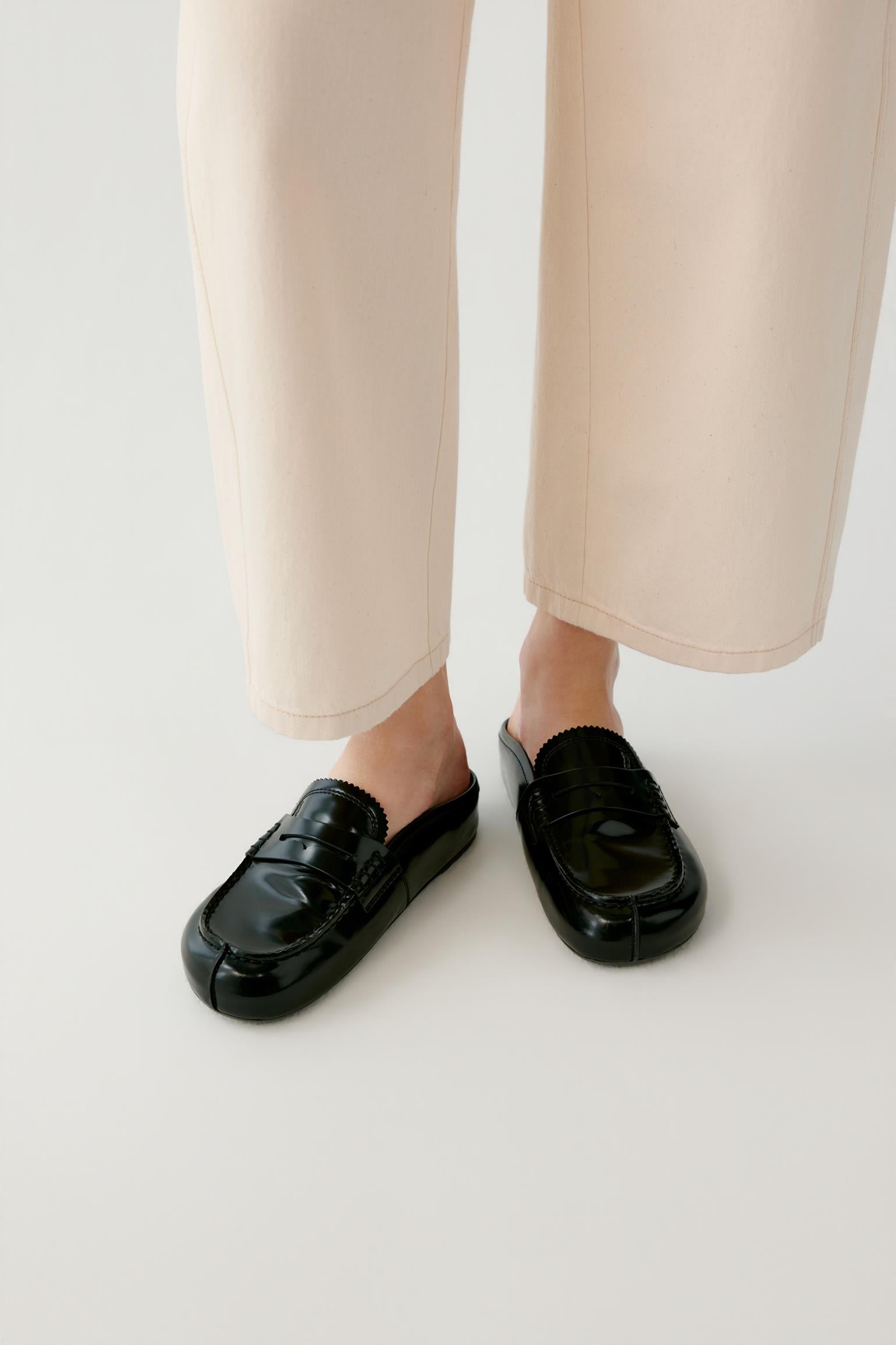 A person wearing black patent leather penny loafers with a unique V-shaped toe and thick platform soles, paired with light beige wide-leg trousers, against a plain light gray background.