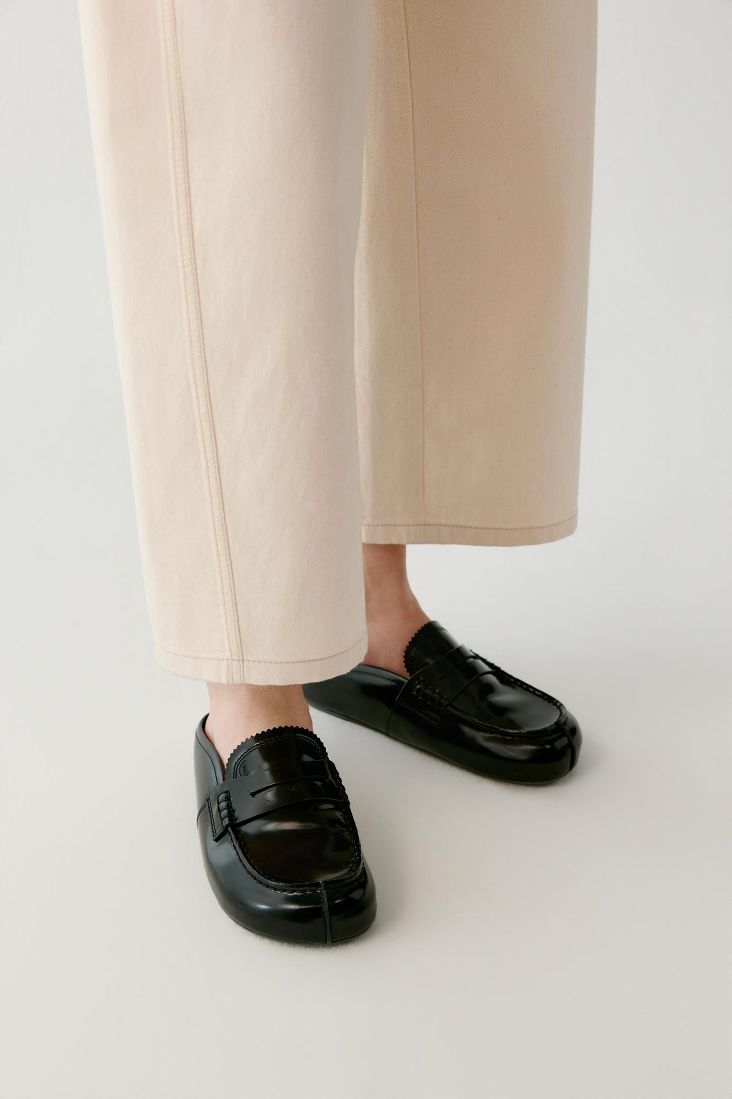 A close-up of a person's feet wearing black leather penny loafers with a split toe detail, paired with light beige cropped trousers, against a plain light gray background.