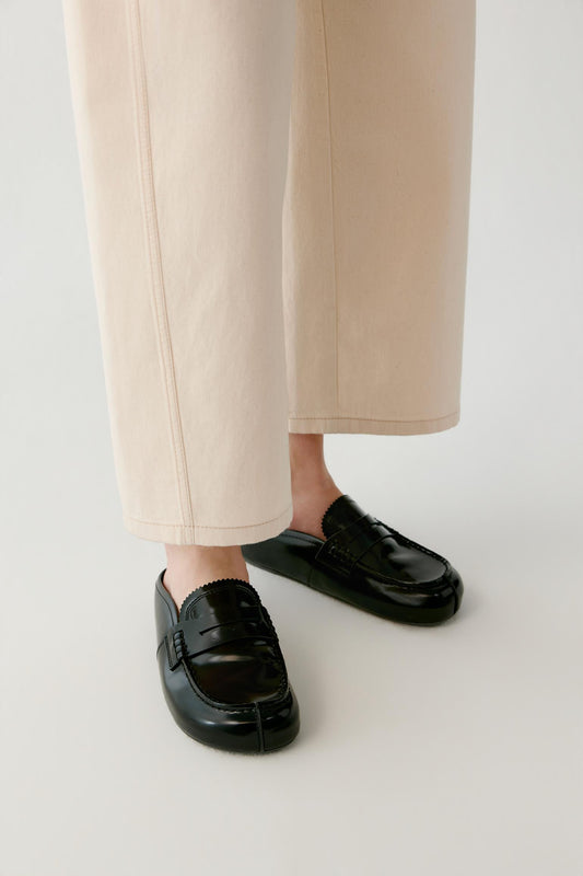 A close-up of a person's feet wearing black leather penny loafers with a split toe detail, paired with light beige cropped trousers, against a plain light gray background.