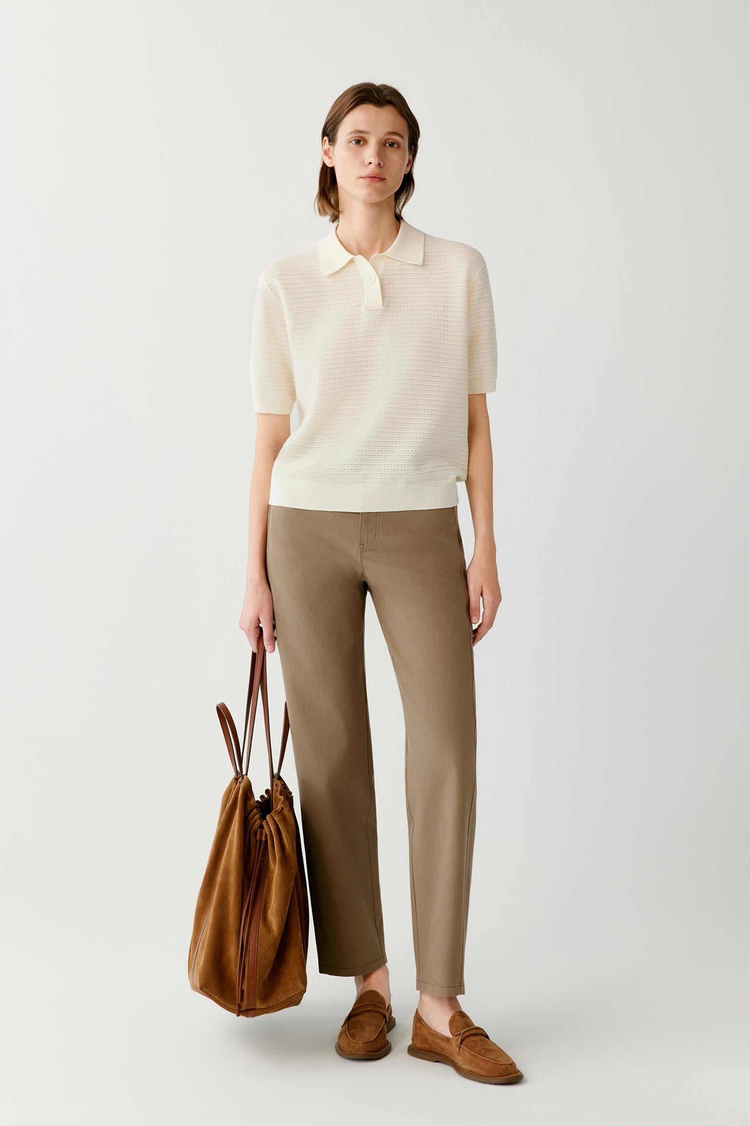 A model stands against a light gray studio background wearing a cream short-sleeve polo knit top, taupe straight-leg trousers, and brown suede loafers, while holding a brown leather tote bag.
